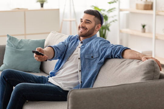 Leisure And Entertainment Concept. Smiling jewish guy watching TV show or film, holding remote control. Young man in kippa enjoying free time sitting on the comfortable couch at home in living room