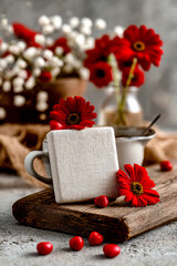 White square cushion leans against coffee cup with red gerbera flowers on wooden board.