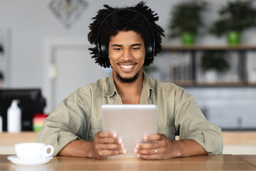 Happy millennial black curly guy in headphones looking at tablet, surfing in internet, sitting at sits at table with cup of coffee in cafe interior. Device, relax with favorite music, funny video