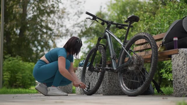 woman crouched fixing bicycle tire, park bench and backpack nearby, close focus on hands and wheel, patching puncture, casual outfit, soft natural light, leafy background, practical urban maintenance