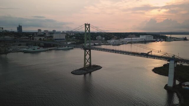 Aerial Footage Shows The Angus L. Macdonald Bridge In Nova Scotia Canada At Summer Sunset With Warm Light Reflecting On Harbor As The Bridge Connects Halifax And Dartmouth Across Calm Coastal Waters