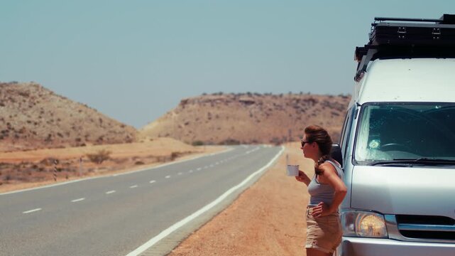 Young adult woman leans against a van on a long, empty road surrounded by brown hills while holding a cup