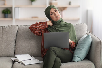 Neckache. Mature Muslim Businesswoman Touching Neck Having Pain After Work On Laptop Computer Sitting On Couch At Home, Wearing Hijab. Health Problems In Older Age Concept