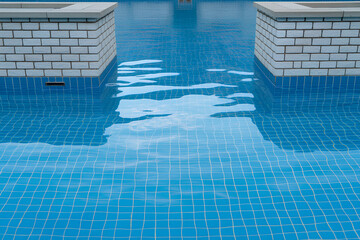 Close up of a blue tiled swimming pool with brick pillars and undulating water surface reflecting sky and clouds