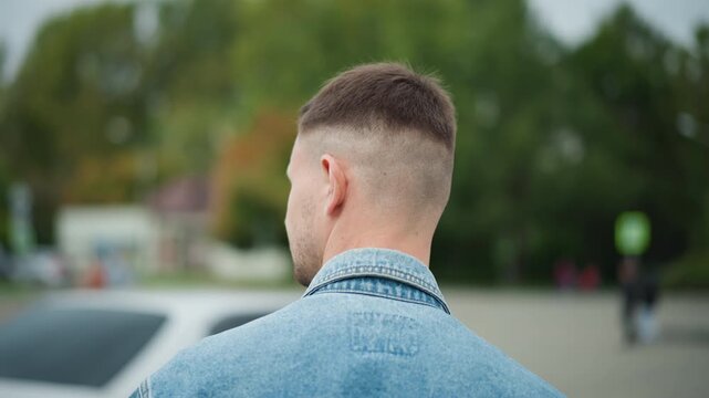 man back portrait facing empty lot, short haircut and denim jacket, slow turn to open car door and enter, closeup shoulder and head shot, intimate anticipation and quiet suburban greenery