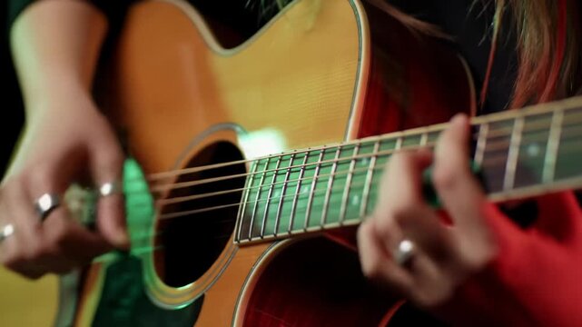 Extreme close-up video of musician's calloused hands expertly playing a glossy acoustic guitar with emerald green and ruby red stage lights, shallow bokeh, cinematic music performance concept.