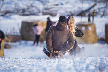 A joyful winter moment showing a man and a woman from behind as they sled down a snow-covered slope together. The man is wearing a brown fleece jacket and a black beanie, while snow spray kicks up aro © Roland