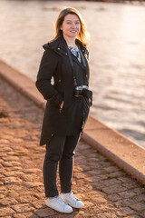 Young Woman Standing Outdoors with a Camera by the Water during Sunset. 