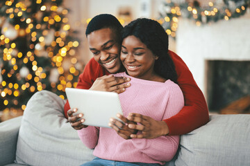 Naklejka premium A couple sits together on a gray sofa, smiling and looking at a tablet. The room is decorated for the holiday season with lights and a Christmas tree in the background.