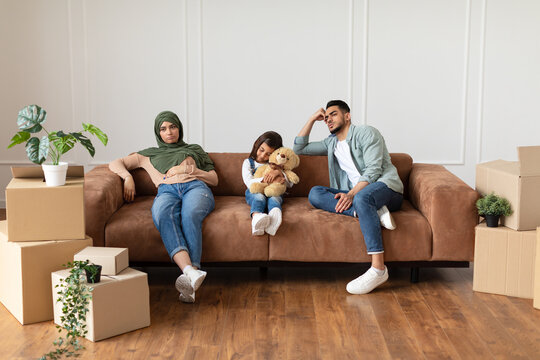 Portrait of sad and tired muslim family of three people sitting on the couch in living room with cardboard boxes around. Exhausted dad, mom in headscarf and girl with teddy bear unpacking things