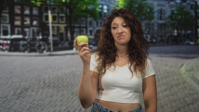 Woman holding a green apple on a cobblestone street, grimacing with hand near chest, wearing white crop top and jeans against blurred buildings; disgust reluctance.