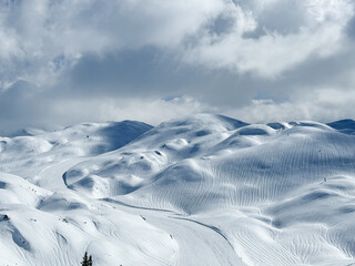 Rugged alpine landscape covered in textured snow beneath a dramatic winter sky. Subtle mountain ridges and smooth contours are interspersed with natural patterns shaped by early spring rain and wind.