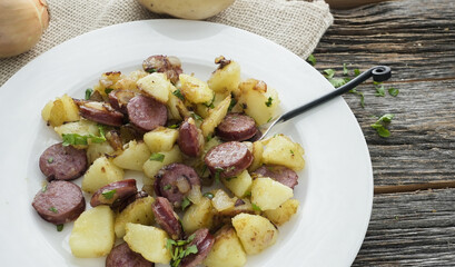 Dish of fried potatoes and sausages served on a plate on wooden table with herbs and utensils