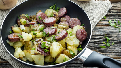 Sausage and potatoes are cooking together in a frying pan. The dish looks fresh with green herbs on top. The setting includes a wooden table and a textured cloth.