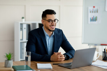 Fototapeta premium Confident young Arab entrepreneur in formal suit using laptop computer at his desk, having virtual meeting at office. Millennial businessman using pc for online job, communicating remotely