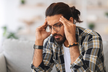 Closeup of sad arab guy sitting on couch in living room, touching his head, suffering from...