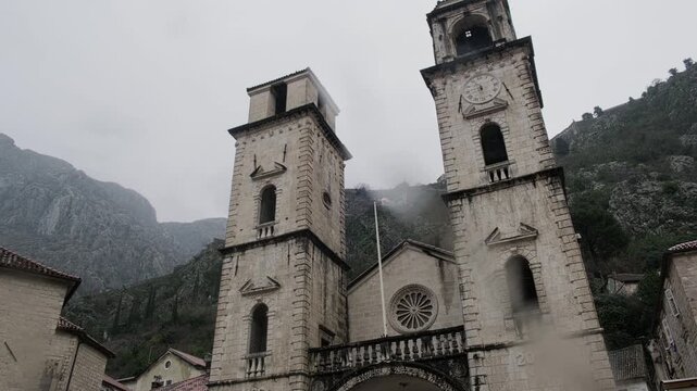 Low-angle view of historic stone cathedral with dual bell towers set against misty mountains on overcast rainy day. The Cathedral of Saint Tryphon in Kotor, Montenegro