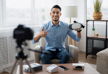 Trendy Arab influencer filming brand advertising promo, presenting VR headset at camera from home. Happy male vlogger recording video about innovative device for technology blog. Tech product review