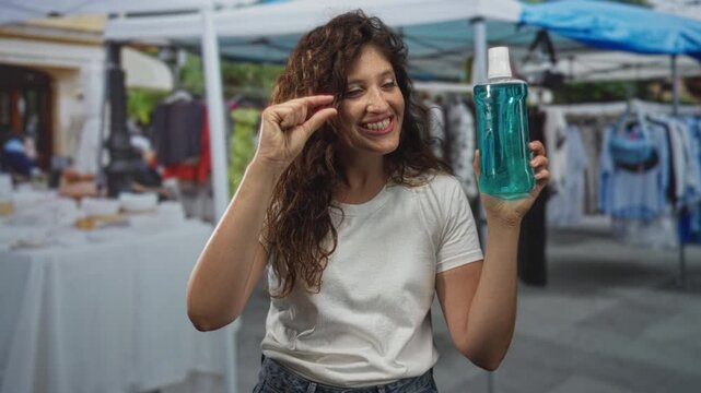 Woman holds a blue bottle and pinches thumb and forefinger at a street market stall to show size; cheerful product demo.