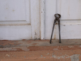 Old forged pincers standing on wooden doorstep near worn white doors.