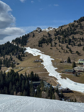 Alpine ski slope winds down a mountainside toward small buildings as winter snow gives way to early spring terrain. Patches of snow contrast with brown hills and trees. Seasonal transition in the Alps