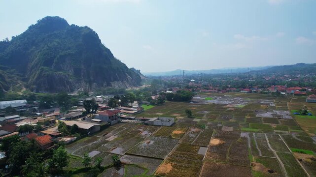 Aerial drone footage of many muddy rectangle rice fields nearby a main road and a village, with Gunung Kuda mountain behind, with a stone quarry, in Majalengka regency, Java island, Indonesia 