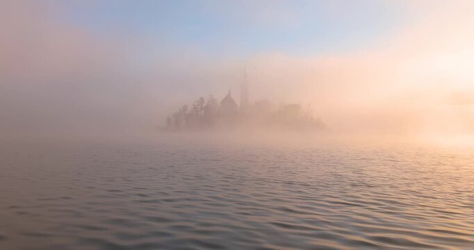 Die Kirche auf der Insel im See Bled im Morgengrauen eingerahmt von gold gef&auml;rbtem Nebel im warmen Licht der goldenen Stunde vor bewaldetem Ufer und ruhigem Wasser