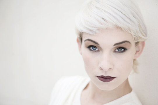 A female model with short blond hair looks towards the camera. She is wearing a cream dress and dramatic makeup.