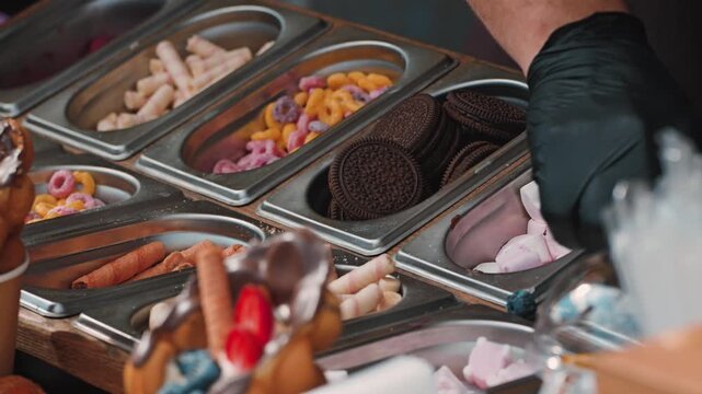A variety of colorful toppings for desserts are displayed in stainless steel trays, including chocolate cookies, marshmallows, and fruity candies. A gloved hand is shown preparing a sweet treat