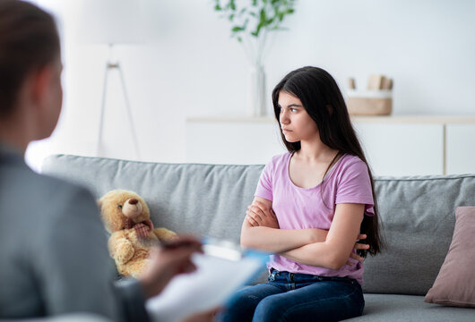 Unhappy Indian teen girl having session with psychologist at clinic, sitting with crossed arms, thinking about problems. Counselor speaking with teenage patient at office, offering help and support