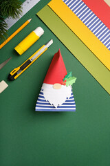 Work desk with sheets of paper, glue, ruler, and eraser on green background. Finished paper craft gnome