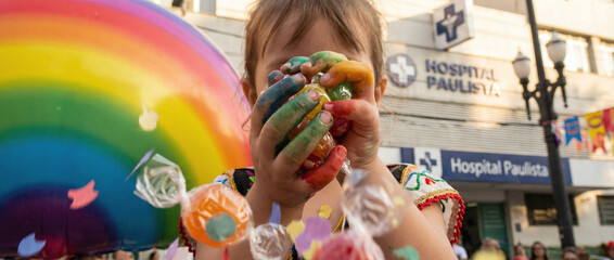 a young girl with brown hair, wearing a colorful dress, holding rainbow-smeared hands over face, with a large rainbow balloon and colorful lollipops in the background.