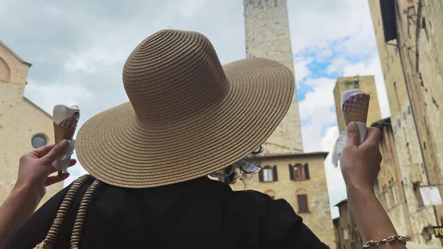 Stylish woman wearing a wide-brimmed hat holds two gelato ice creams cones while exploring the historic town of San Gimignano, Tuscany