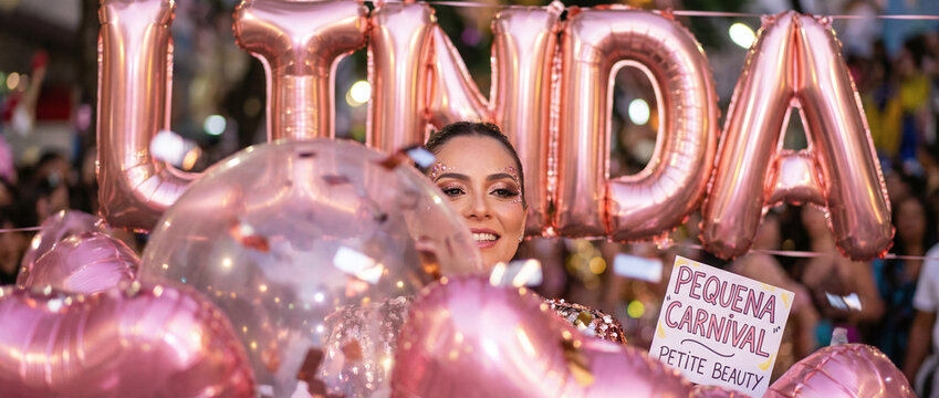 a smiling woman with dark hair, surrounded by pink balloons and "UMA" letters, holding a "Felicita Carnevale Petto Reale" sign, in a festive, brightly lit crowd.