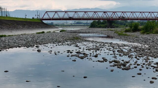 A bridge spans a river with a rocky shoreline. The bridge is red and spans the entire width of the river. The water is calm and the rocks on the shoreline are scattered