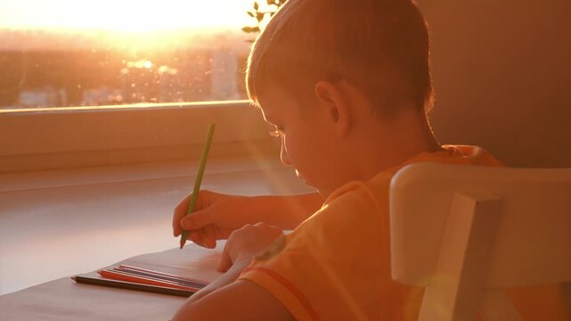 Focused young student. Young boy concentrates attentively on his schoolwork amid sunlight and city view. Young child carefully drawing colorful pencils while sunlight illuminates room and city skyline