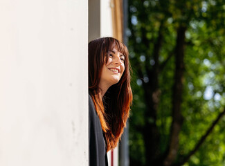 Fototapeta premium Smiling woman with long brown hair leans out of a window, enjoying the sunlight and greenery outside, creating a cheerful and vibrant atmosphere of happiness and joy