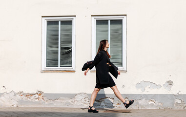 Obraz premium Young woman in black dress and sandals walks confidently along a textured wall, showcasing urban style and modern fashion in a vibrant city environment