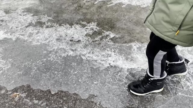 A child is walking on a snowy and icy walkway, wearing winter boots and warm clothing. The child is stomping on the ice thereby breaking it. The icy surface contrasts with the colorful clothing.
