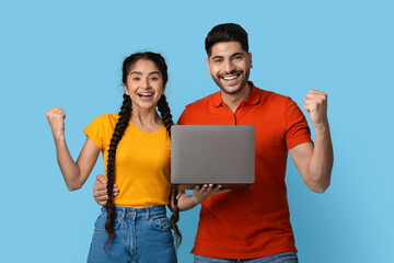 A cheerful young man and woman stand together, smiling widely and celebrating. They hold a laptop, showing their excitement for an achievement. The background is bright blue.