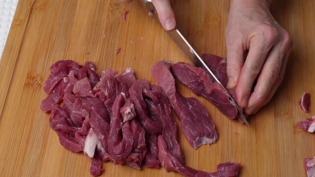 Slicing beef into thin strips on wooden board. Hands slicing raw boneless beef into long thin strips on a wooden cutting board. Close-up meat preparation for cooking.