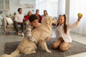 Cute children playing with their golden retriever on floor at home, their relatives sitting on couch on background. Adorable kids having fun with pet dog during family gathering, indoors
