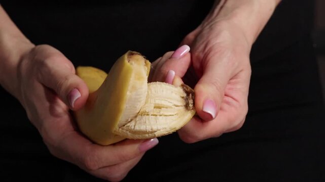 Person peels a banana in a close-up shot showing hands and fruit details during a casual moment. Woman's hands hold a banana and peels it with both hands