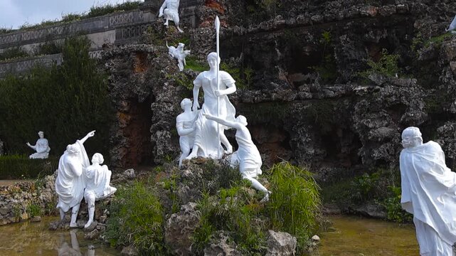 Detailed view of white classical statues in a rocky grotto fountain at Quinta Real de Caxias, surrounded by greenery and shallow water in a historic garden setting.
