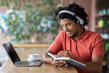 Happy Young Black Guy Taking Notes To Notepad While Studying Online With Laptop In Cafe, Millennial African Man In Wireless Headphones Noting Information From Webinar, Enjoying Remote Education