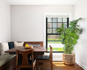 A cozy dining room with a wood table surrounded by wood chairs and built in bench. A bright window shines light on a plant and hardwood flooring © Joseph Hendrickson