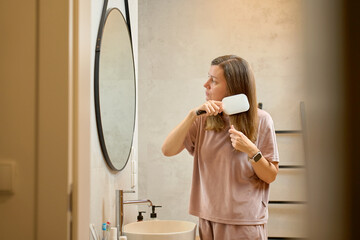 Woman standing in bathroom and brushing hair while looking at mirror
