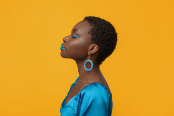 Profile portrait of stylish short-haired millennial black woman with closed eyes posing on yellow studio background, wearing bright accessories and blue makeup, copy space © Prostock-studio