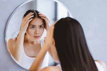 A woman stands in front of a round mirror looking at herself. She holds her hair back with both...