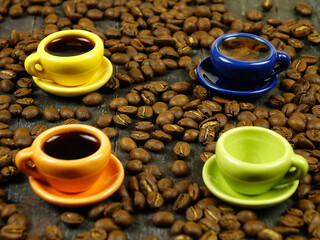 Small colorful cups with hot black coffe among scattered coffee beans are on a black wooden board, side view
